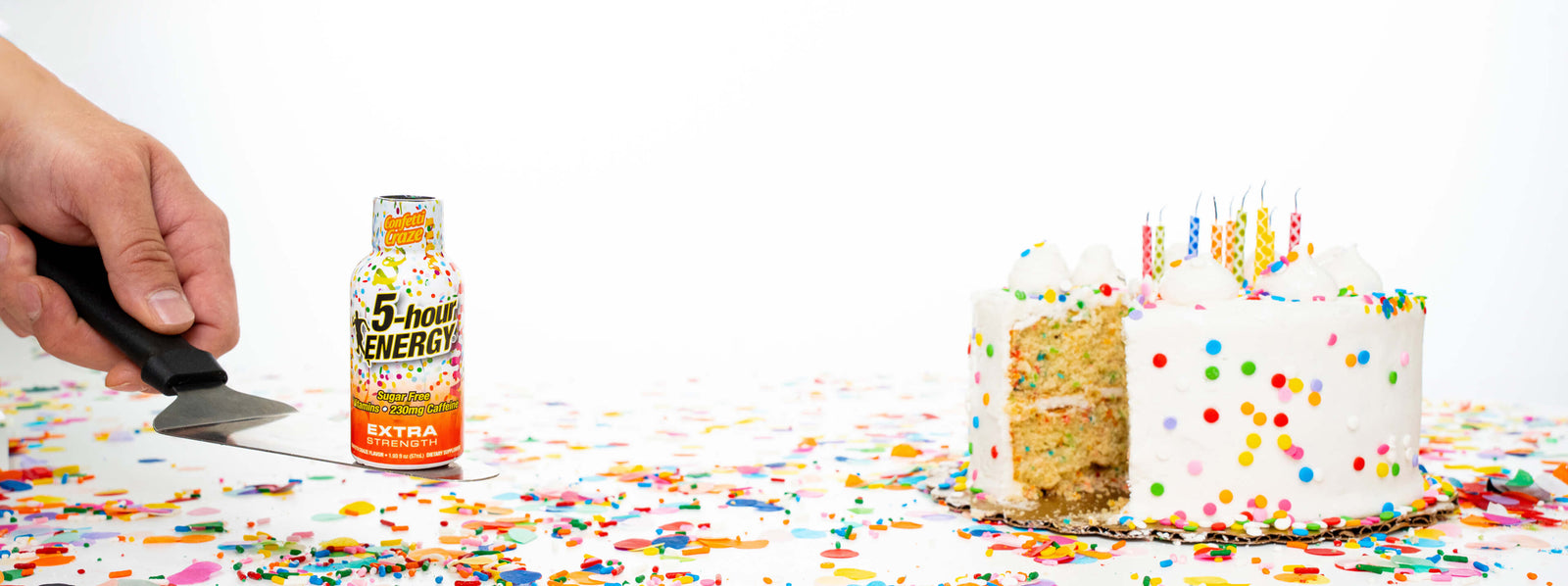Hand holding a bottle labeled '5-hour Energy' next to a birthday cake with colorful sprinkles on a white background.