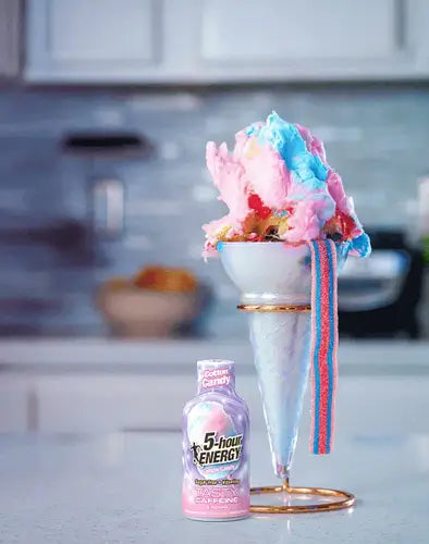 A pink bottle of Cotton Candy 5-hour ENERGY® next to a tall, decorated ice cream sundae in a glass cone, set on a kitchen counter with a blurred kitchen background.