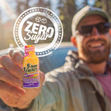 A man in sunglasses and a cap holds up a small bottle of 5-hour Energy Tasty Caffeine Energy Drink in front of a blurred outdoor background. A large circular graphic overlay reads 'ZERO Sugar' and partially covers the bottle.