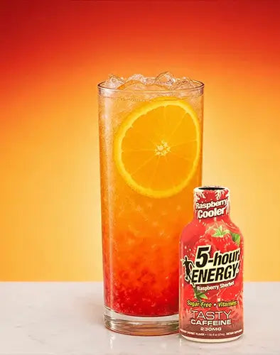 A tall glass of 5-hour Energy Cooler raspberry lemonade iced coffee with a fresh orange slice, shown next to a bottle of the beverage on a white surface against a vibrant orange gradient background.