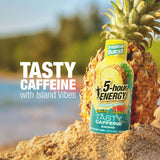 A bottle of Tropical Burst 5-hour Energy Pineapple-Apricot flavored caffeine drink stands upright in sand next to a whole pineapple, with blurred beach scenery in the background and large text reading 'TASTY CAFFEINE with Island Vibes'.