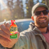 A man in sunglasses and a cap holds up a small bottle of 5-hour Energy drink to the camera, with a blurred outdoor background and a car visible behind him.