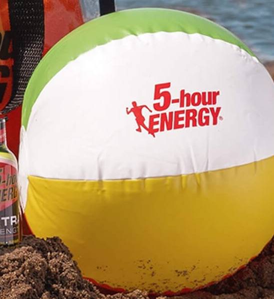 Beach ball with 5-hour ENERGY logo on a sandy surface next to a bottle of 5-hour ENERGY drink.