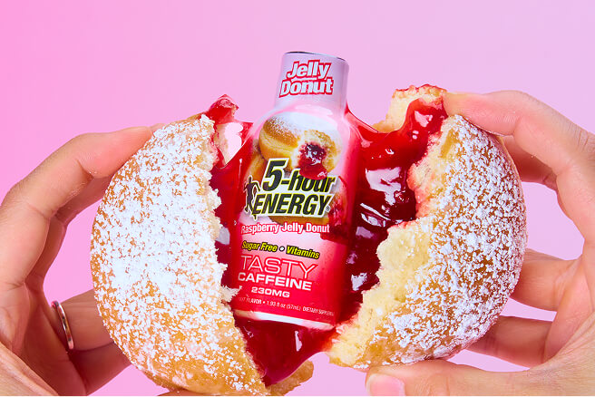 Two hands hold a powdered jelly donut split open to reveal a red jelly filling and a 5-hour Energy drink bottle embedded in the center, against a solid pink background.