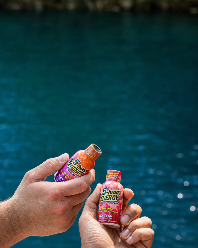 Two hands holding two bottles of 5 Hour Energy drink, one orange and one red, with a blurred blue water background.