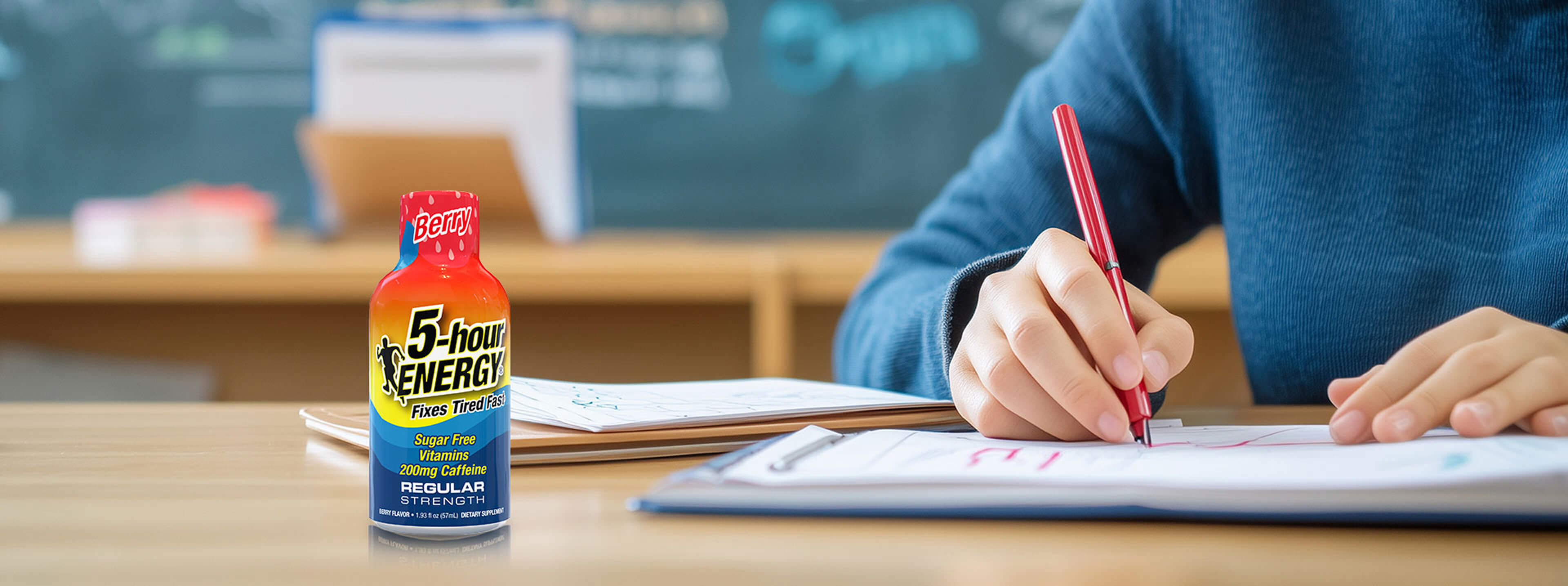 Person writing with a red pen next to a bottle of 5-hour Energy drink on a desk.