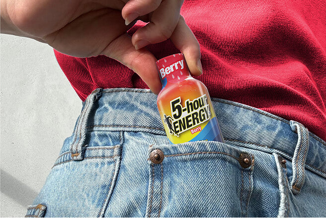 A person wearing a red shirt and light blue denim jeans holds a small bottle of Berry 5-hour Energy drink tucked into the back pocket of the jeans.